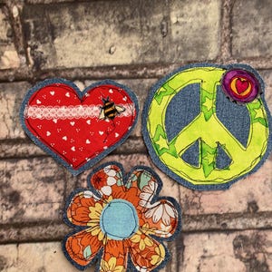 May include: Three denim-edged fabric patches: a red heart with a bee, a yellow peace sign, and an orange flower. The heart has a white lace trim and small white hearts. The peace sign has a purple flower with a heart center. The flower has a blue center.