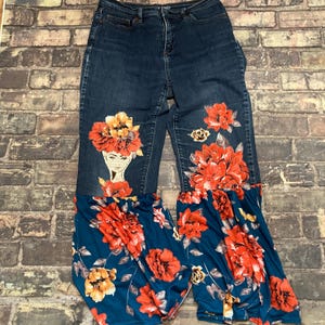 May include: Dark blue denim jeans featuring a high-waisted fit with a zipper and button closure. The upper legs display a printed graphic of a face adorned with orange flowers, while the lower legs transition into flared floral fabric.