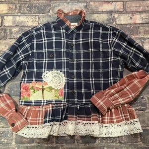 May include: A long-sleeve button-up shirt featuring a navy blue and white plaid pattern. The shirt includes a red and white plaid collar, cuffs, and hem, accented with a white lace trim, a round white doily, and a rectangular floral patch with a dragonfly.
