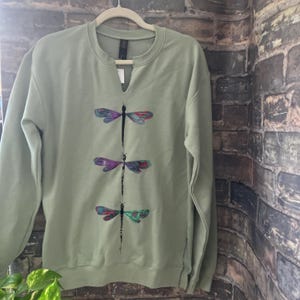 May include: A sage green long-sleeve sweatshirt featuring a notched neckline and a vertical graphic print of three dragonflies. Each dragonfly has a black body and wings patterned with shades of purple, blue, and red. The garment has long sleeves.