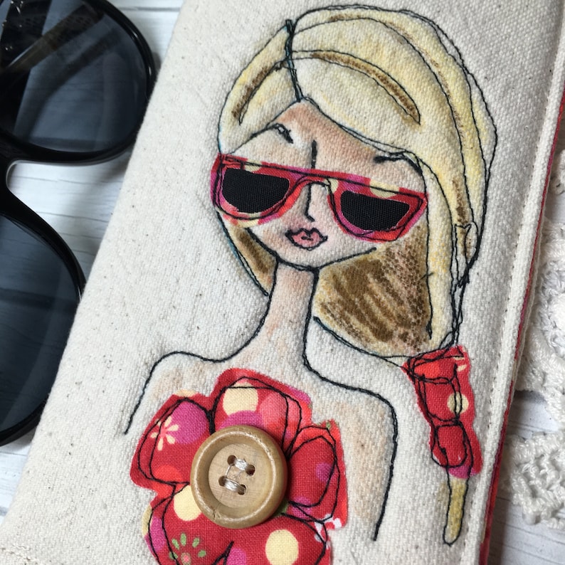 Op de afbeelding: Een wit canvas zonnebrillenetui met een handgestikt ontwerp van een vrouw met blond haar die een rode zonnebril draagt en een rode bloem met een bruine knoop in het midden.