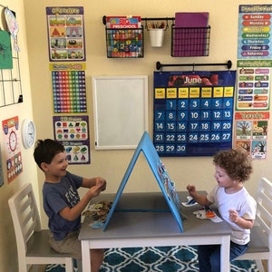 May include: Two children sit at a white table with a blue easel in the middle. The easel has a white board on one side and a blue felt board on the other. The children are playing with colorful felt pieces. The room is decorated with educational posters, a calendar, and a clock.