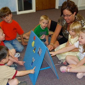 May include: A group of children and an adult are gathered around a blue felt board with colorful picture magnets. The children are pointing at the board, engaged in an educational activity. The setting appears to be a classroom or learning environment.