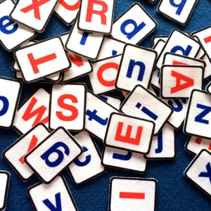 May include: A collection of white letter tiles with black borders, each featuring a single letter in red or blue. The letters are scattered on a blue surface.