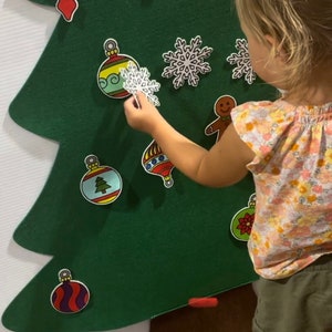 May include: A felt Christmas tree with various ornaments, including a star, candy canes, snowflakes, and ornaments. A child is placing a snowflake ornament on the tree.
