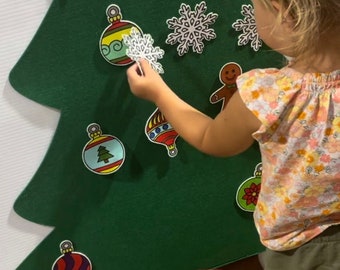 Puede incluir: Un árbol de Navidad de fieltro con varios adornos, incluyendo una estrella, bastones de caramelo, copos de nieve y bolas. Un niño está colocando un adorno de copo de nieve en el árbol.