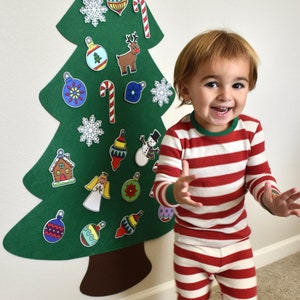 May include: A child wearing red and white striped pajamas stands in front of a felt Christmas tree with various ornaments and decorations. The tree is green and has a brown trunk. The child is smiling and looking at the camera.