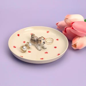 May include: A white ceramic trinket dish with a red heart pattern. A small ceramic dog figurine is in the centre of the dish. There are two pairs of earrings and a ring on the dish.