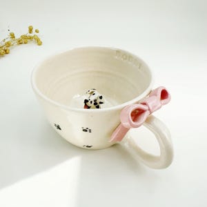 May include: Cream-colored ceramic mug with a pink bow handle and paw print accents. A small Dalmatian dog figurine is inside the mug. The word "Dolly" is written on the rim.