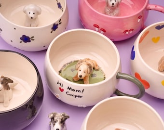 May include: A collection of ceramic mugs with dog figurines inside. The mugs are various colors, including white, pink, and black, and are decorated with floral and mushroom designs. One mug has the text "Hi Mom! Cooper".