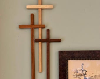 May include: A wooden cross wall sculpture with four crosses in shades of brown, from light to dark. The crosses are layered and overlapping, creating a textured effect. The artwork is mounted on a pale green wall, adding a touch of colour.