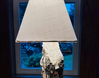May include: A table lamp featuring a base made from a natural birch log segment with white and black bark, mounted on a circular wooden disc. The lamp is topped with a trapezoidal, light beige fabric lampshade with a fine, woven texture.