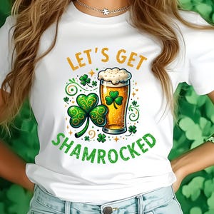 May include: White t-shirt with a green shamrock and a glass of beer. The text "Let's Get Shamrocked" is printed on the shirt.