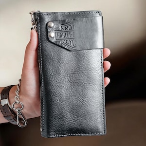 Può includere: Un portafoglio in pelle nera con chiusura a zip e catena metallica. Il portafoglio presenta il logo Harley-Davidson in rilievo sul davanti. Il portafoglio è tenuto in mano da una persona.