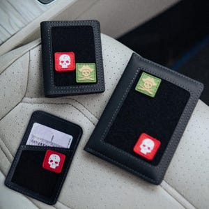 May include: Three black leather wallets with skull and crossbones patches. One wallet has a card visible. The patches are red and green with white skull designs. The wallets are on a light-coloured surface.