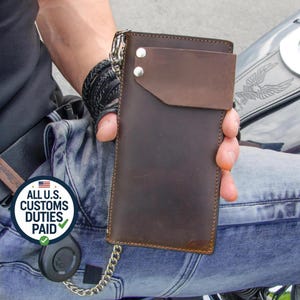 Op de afbeelding: Een bruine leren portemonnee met een flap en zilveren ketting. De portemonnee heeft een rechthoekige vorm met een donkerbruine flap die is vastgezet met twee zilveren klinknagels. De portemonnee wordt vastgehouden door een persoon die een blauwe spijkerbroek draagt.