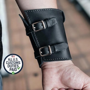 Può includere: Polsino in pelle nera con due fibbie color argento. Il polsino è cucito con filo bianco e ha una forma rettangolare. L'immagine include anche una grafica circolare con il testo "ALL U.S. CUSTOMS DUTIES PAID".