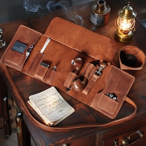 Puede incluir: Un estuche de cuero marrón para pipa de tabaco, abierto sobre una superficie de madera, con una pipa, un encendedor y otros accesorios. También se ven una linterna vintage y un pequeño cuenco de cuero. Una hoja de papel con texto manuscrito está en primer plano.
