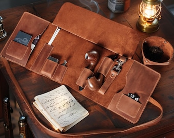 May include: A tan leather tobacco pipe roll featuring multiple interior pockets and slots. The roll contains a wooden tobacco pipe, a metal tool, and a small black card. A stack of handwritten notes rests on the leather surface of the open organizer.