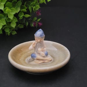 May include: A small ceramic figurine of a gnome wearing a blue hat and sitting on a brown and white ceramic dish.