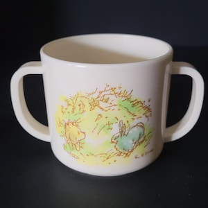 May include: White plastic cup with two handles featuring a Winnie the Pooh illustration of Pooh Bear and Rabbit in a green field with yellow flowers. The text "© Disney" is printed on the bottom of the cup.