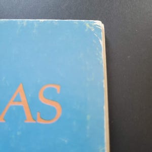 K&ouml;nnte beinhalten: Nahaufnahme eines Buchcovers mit den orangefarbenen Buchstaben "AS" auf hellblauem Hintergrund. Die Kanten des Buches sind sichtbar und weisen Abnutzungserscheinungen auf. Der Hintergrund ist dunkelgrau.