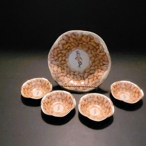 May include: A set of five peanut-shaped ceramic dishes featuring a cartoon peanut character. The larger dish has a rim of gold paint and the smaller dishes have a gold rim and a gold base.