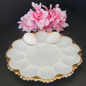 May include: White ceramic egg plate with a gold rim and eight individual indentations for holding eggs. Two white eggs are in the plate.