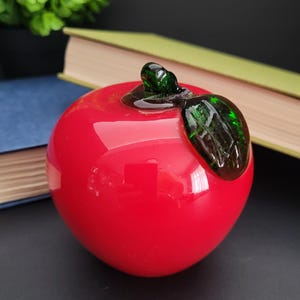 May include: A shiny red glass apple with a green stem and leaf. The decorative fruit is set against a blurred background with books and a plant. The apple is a vibrant, eye-catching piece.