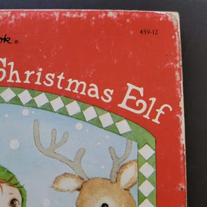 K&ouml;nnte beinhalten: Ein Vintage-Kinderbuch mit dem Titel "Christmas Elf" mit rotem Einband. Das Cover zeigt eine Illustration eines Rentiers und eines Elfen. Der Titel des Buches ist in Wei&szlig; gehalten, mit einem gr&uuml;n-wei&szlig;en Rautenmuster.