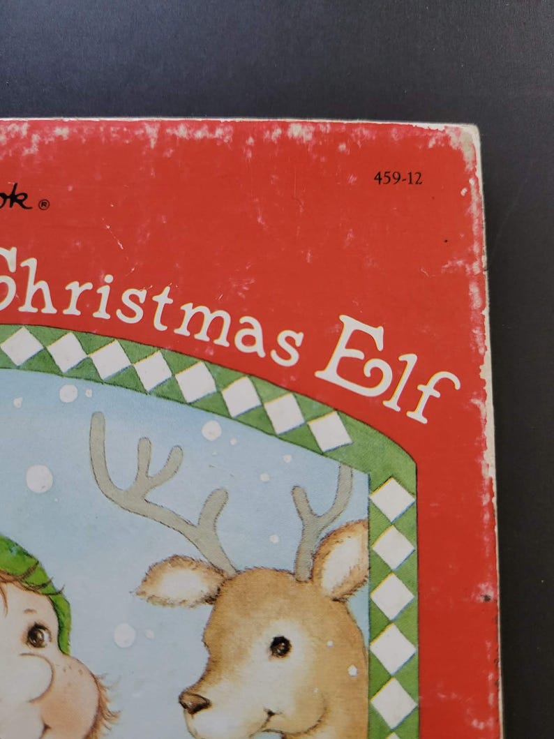 K&ouml;nnte beinhalten: Ein Vintage-Kinderbuch mit dem Titel "Christmas Elf" mit rotem Einband. Das Cover zeigt eine Illustration eines Rentiers und eines Elfen. Der Titel des Buches ist in Wei&szlig; gehalten, mit einem gr&uuml;n-wei&szlig;en Rautenmuster.