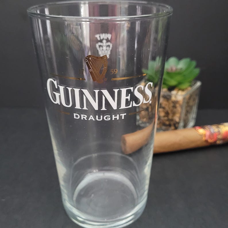 May include: A clear glass pint glass with the Guinness logo in gold and white. The logo includes the text "GUINNESS DRAUGHT" and a harp with a crown above it.