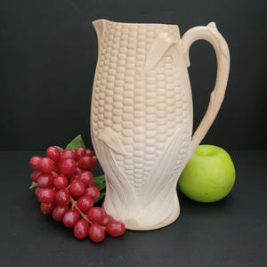 May include: A white ceramic jug shaped like an ear of corn. The jug has a handle and a textured surface.