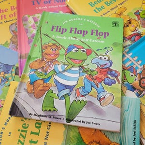 May include: A selection of children's books featuring Jim Henson's Muppets. The central book, "Flip Flap Flop," has a green cover with Kermit the Frog and other characters. Other books include titles such as "The Best Gift of..." and "TV or Not TV."