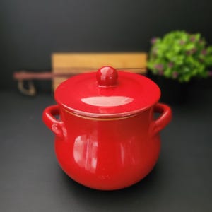 May include: A small, round, red ceramic cooking pot with a matching lid and two side handles. The lid has a small knob on top. The pot is glossy and sits on a dark surface. A decorative item for the kitchen.