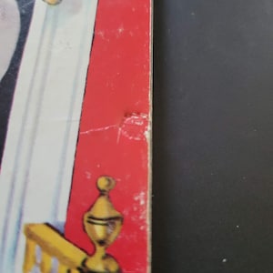 K&ouml;nnte beinhalten: Ein Abschnitt eines Vintage-Buchcovers mit rotem Buchr&uuml;cken und einem Teil einer gelb-wei&szlig;en Illustration. Der Buchr&uuml;cken weist einige Abnutzungserscheinungen auf. Die Illustration zeigt ein goldfarbenes dekoratives Element.