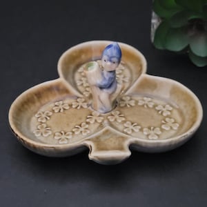 May include: A brown and white ceramic clover-shaped dish with three sections. A small blue and white figurine of a leprechaun holding a pot of gold sits in the centre of the dish.