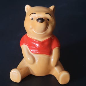 May include: A ceramic Winnie the Pooh figurine. The figurine is sitting down with its arms crossed and is wearing a red shirt. The figurine is brown and has a black nose and eyes.