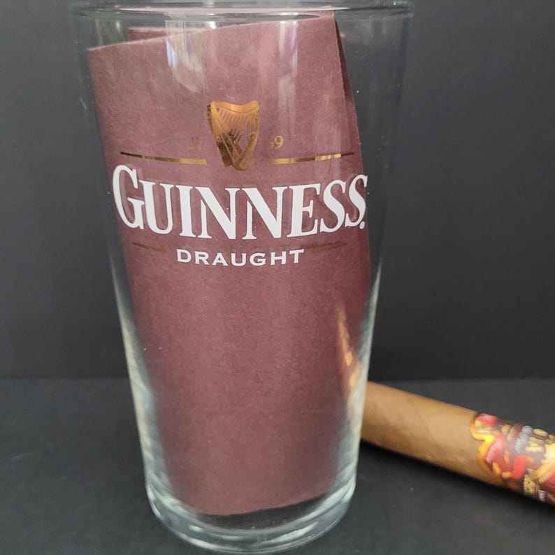 May include: A clear glass pint glass with a gold Guinness logo and the words "Draught" printed on it. A brown cigar is lying on a dark surface next to the glass.