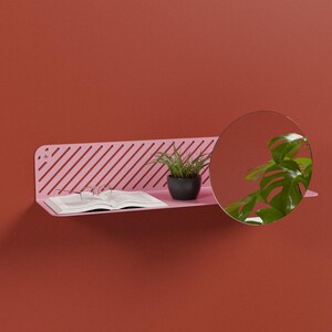 Peut inclure: Une étagère murale en métal rose avec un panneau arrière à lattes diagonales et un miroir circulaire intégré sur le côté droit. L'étagère supporte un livre ouvert aux pages blanches et une petite plante verte dans un pot gris foncé.