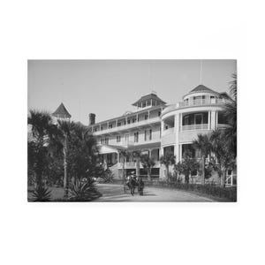 May include: Black and white photograph of a large, multi-story hotel with a circular balcony and multiple turrets. Palm trees line the driveway leading to the entrance. A horse-drawn carriage is visible on the road.