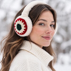 May include: Plush white faux fur earmuffs featuring a circular crochet accent on the side. The crochet detail consists of a dark green center surrounded by a ring of cream-colored petals, all framed by a vibrant red border on a white base.