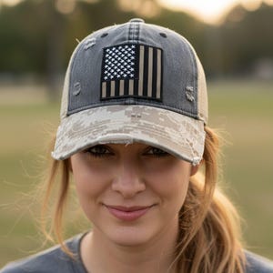 May include: A distressed gray baseball cap with a mesh back and a camouflage brim. The cap features a patch with a black and white American flag design. The hat has a worn, vintage look.