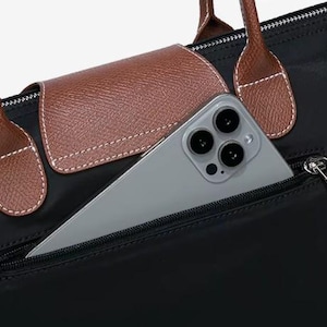 May include: Black tote bag with brown leather accents, including handles and a flap. A silver smartphone is partially visible in the front zippered pocket. The bag has a classic, minimalist design.