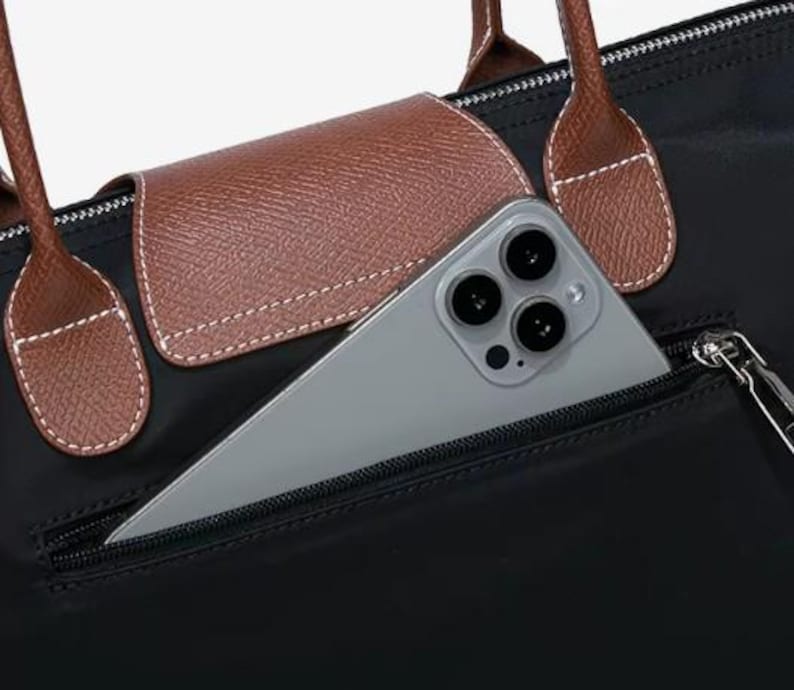 May include: Black tote bag with brown leather accents, including handles and a flap. A silver smartphone is partially visible in the front zippered pocket. The bag has a classic, minimalist design.