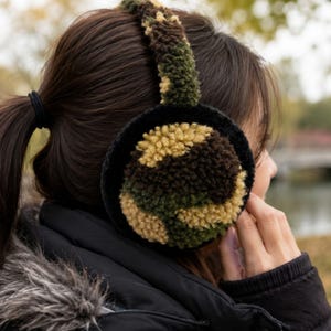 May include: Earmuffs featuring a textured, fuzzy camouflage pattern in shades of dark green, brown, and tan. The circular ear covers are bordered by a solid black trim, and the connecting headband is covered in the same camouflage material.