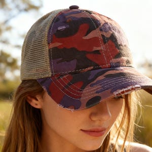 May include: A camouflage baseball cap with a mesh back. The cap features a distressed brim and a pattern of red, purple, and black shapes. The mesh back is tan. The cap is worn by a person.