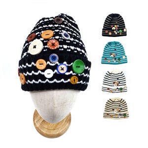 May include: A black and white striped knit beanie adorned with various colorful buttons. The hat is displayed on a mannequin head, showcasing its design. Additional beanies in different colors and button arrangements are also shown.