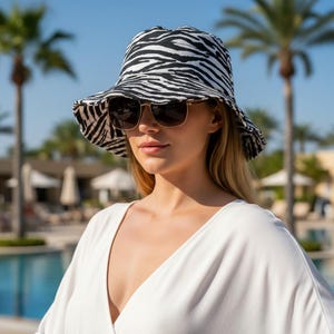 May include: A black and white zebra print bucket hat with a wide brim. The hat is worn with sunglasses and a white top. The background includes palm trees and a swimming pool.