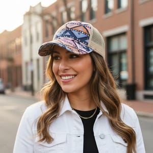 May include: A trucker hat with a patterned design in shades of blue, pink, and orange. The hat has a beige mesh back and a curved brim. The person in the image is wearing a white jacket and a gold necklace.